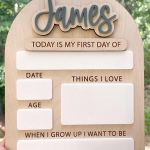 Op de afbeelding: Een boogvormig houten bord met de naam James in verhoogd grijs schrift. Het bord bevat de tekst TODAY IS MY FIRST DAY OF, DATE, THINGS I LOVE, AGE en WHEN I GROW UP I WANT TO BE, met lege witte rechthoekige vakken om op te schrijven.