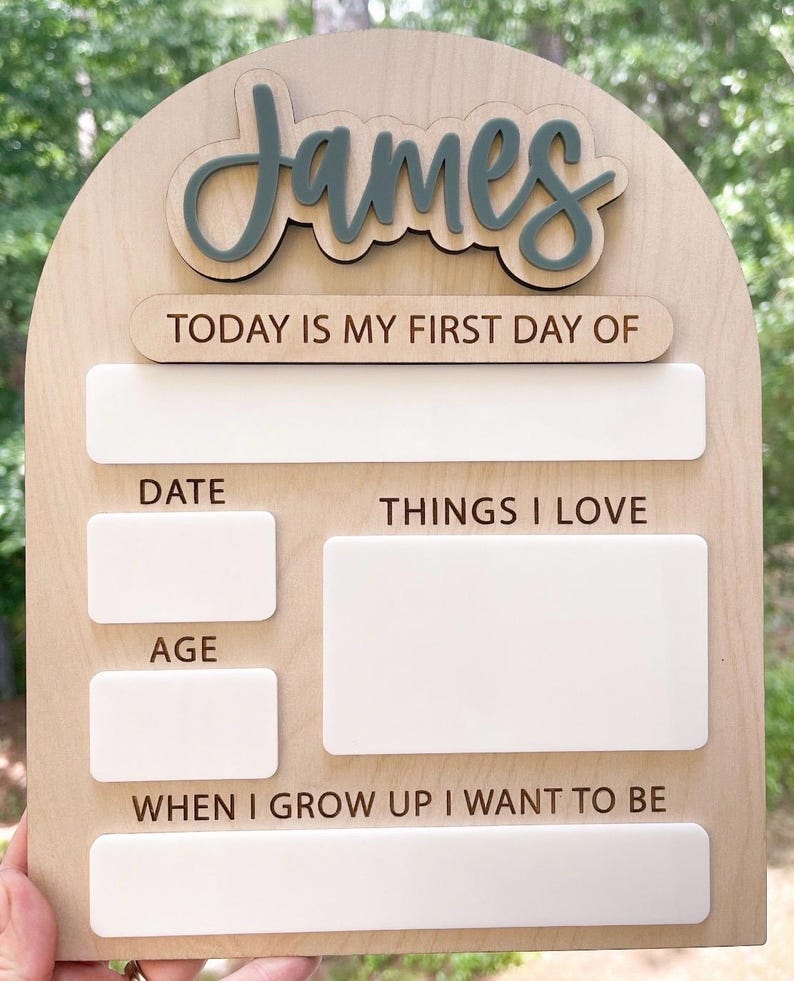 Op de afbeelding: Een boogvormig houten bord met de naam James in verhoogd grijs schrift. Het bord bevat de tekst TODAY IS MY FIRST DAY OF, DATE, THINGS I LOVE, AGE en WHEN I GROW UP I WANT TO BE, met lege witte rechthoekige vakken om op te schrijven.