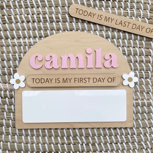 May include: A semi-circular wooden sign featuring the name camila in raised pink letters, the text TODAY IS MY FIRST DAY OF, and a white rectangular space for writing. Two white flowers with gold centers are attached to the sides of the sign.
