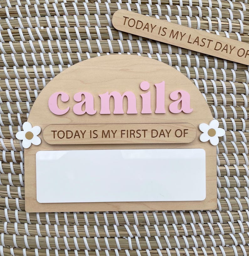 May include: A semi-circular wooden sign featuring the name camila in raised pink letters, the text TODAY IS MY FIRST DAY OF, and a white rectangular space for writing. Two white flowers with gold centers are attached to the sides of the sign.