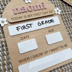 May include: An arched wooden sign featuring the name naomi in pink letters, with white flower accents. It includes the text TODAY IS MY FIRST DAY OF, FIRST GRADE, DATE, AGE, and WHEN I GROW UP I WANT TO BE, with blank white spaces for writing.