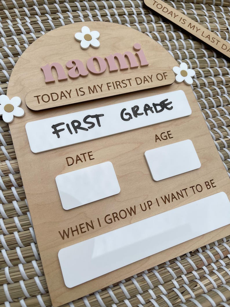May include: An arched wooden sign featuring the name naomi in pink letters, with white flower accents. It includes the text TODAY IS MY FIRST DAY OF, FIRST GRADE, DATE, AGE, and WHEN I GROW UP I WANT TO BE, with blank white spaces for writing.