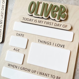 Op de afbeelding: Een houten bord met de naam OLIVER in verhoogde groene letters. Het bord bevat de tekst TODAY IS MY FIRST DAY OF, DATE, AGE, THINGS I LOVE en WHEN I GROW UP I WANT TO BE, met witte rechthoekige vakken om informatie op te schrijven.