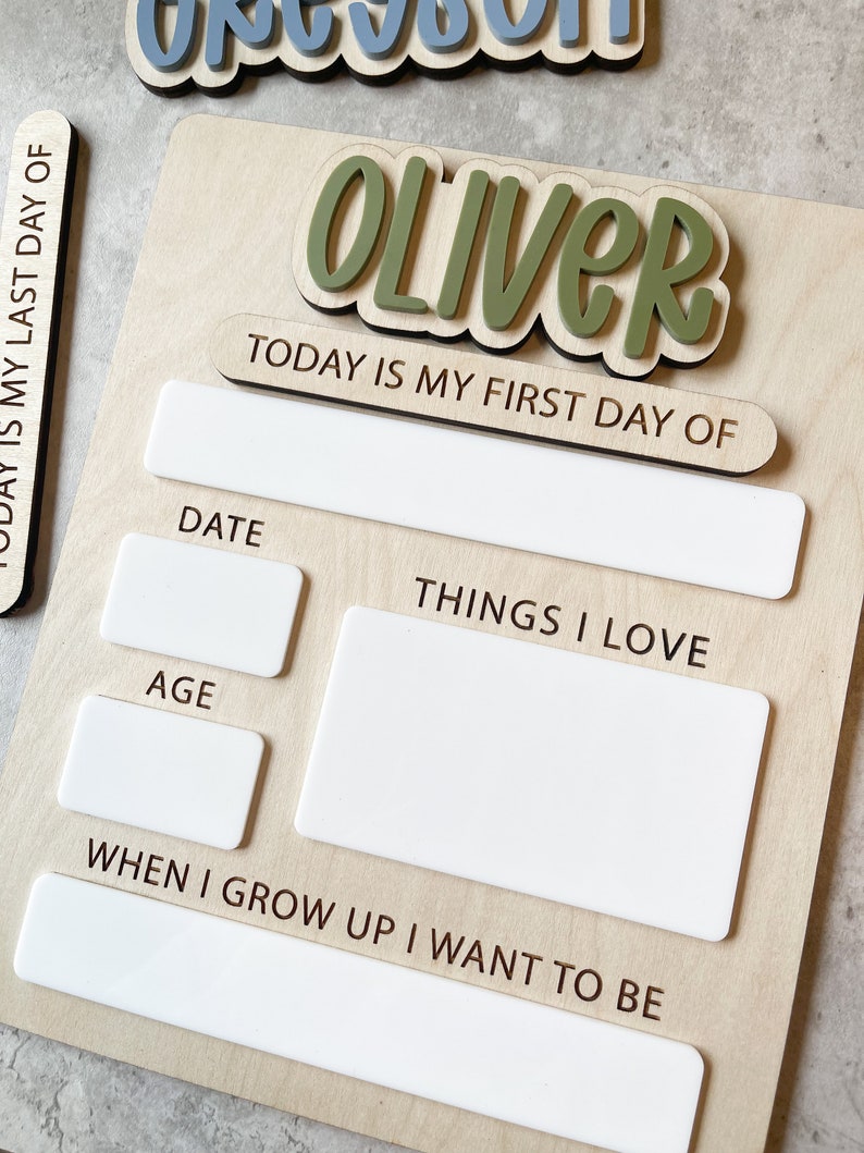 Op de afbeelding: Een houten bord met de naam OLIVER in verhoogde groene letters. Het bord bevat de tekst TODAY IS MY FIRST DAY OF, DATE, AGE, THINGS I LOVE en WHEN I GROW UP I WANT TO BE, met witte rechthoekige vakken om informatie op te schrijven.
