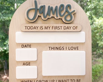 May include: A wooden sign with a light brown background and white accents. The sign is shaped like an arch and has the name "James" written in green cursive letters at the top. The sign also has the text "TODAY IS MY FIRST DAY OF", "DATE", "THINGS I LOVE", "AGE", and "WHEN I GROW UP I WANT TO BE" written on it.