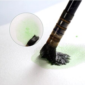 May include: A close-up of a black paintbrush with a green paint stroke on white paper. The brush is being used to create a green watercolor wash.