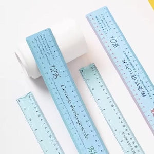 May include: Four light blue rulers with black markings and text, including "12%" and "Ceramic shrinkage scale." The rulers are of varying lengths, with centimeter measurements. One ruler is partially obscuring a white cylindrical object.