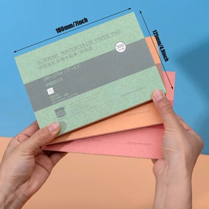 May include: A stack of four watercolor paper pads. The top pad is green and has the text "ACADEMY WATERCOLOR PAPER PAD" in black. The text "中国宝虹全棉水彩本/学院级" is also printed on the pad in black. The pad is 7 inches by 4.9 inches and 300 gsm. The text "180&times;125/7"x4,9" 中粗纹/CP" is printed on the pad in black. The text "120 100%Cotton/Acid Free" is printed on the pad in black. The text "300 G/M" is printed on the pad in black. The text "SAGNONG" is printed on the pad in black.