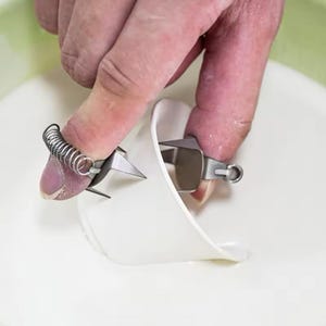 Op de afbeelding: Close-up van een roestvrijstalen gereedschap met een veermechanisme, waarschijnlijk om te snijden of te vormen. Het gereedschap wordt met vingers vastgehouden en boven een wit, gebogen oppervlak geplaatst. Het ontwerp omvat scherpe, schuine messen.