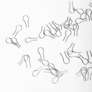 Op de afbeelding: Een verzameling kleine, zilverkleurige splitpennen verspreid op een witte ondergrond. Elke pen heeft een lusvormige kop en twee rechte poten, ontworpen om mechanische onderdelen te bevestigen. De pennen zijn gemaakt van glanzend metaal.