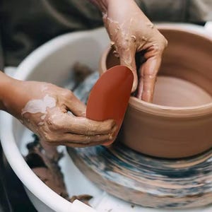 Puede incluir: Unas manos usando una herramienta de cerámica roja para dar forma a una olla de barro en un torno de alfarería.