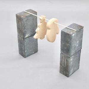 Puede incluir: Dos adornos de madera de color claro, un árbol de Navidad y una seta, cuelgan de una fina clavija de madera. La clavija está sostenida por dos pilas de bloques de hormigón grises, creando una exhibición minimalista.