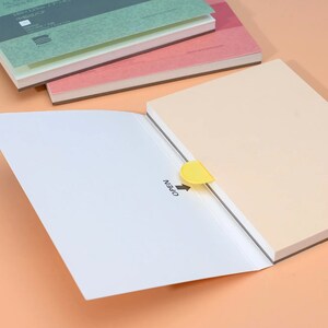 May include: A white notebook with a yellow tab that says "OPEN" is open to reveal a beige notebook. The beige notebook is stacked on top of two other notebooks, one pink and one green.