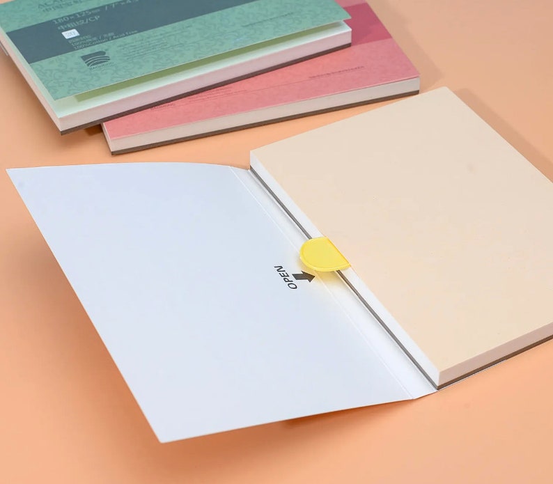 May include: A white notebook with a yellow tab that says "OPEN" is open to reveal a beige notebook. The beige notebook is stacked on top of two other notebooks, one pink and one green.