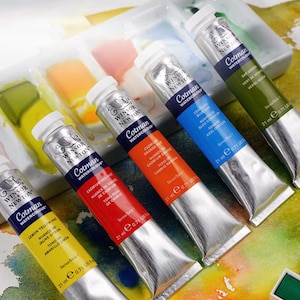 May include: Five tubes of Winsor & Newton Cotman watercolour paint in shades of yellow, red, orange, blue, and green. Each tube contains 21 ml of paint. The tubes are positioned near a white palette with visible paint.