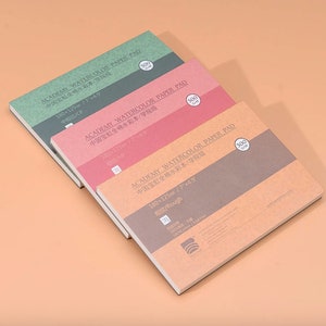 May include: Three watercolor paper pads in different colors and textures. The pads are labeled "ACADEMY WATERCOLOR PAPER PAD" and have the text "中国宝虹全棉水彩本/学院级" in Chinese. The pads are 180 x 125 mm (7 x 4.9 inches) and have different textures: "中粗纹/CP" (medium grain), "细纹/HP" (fine grain), and "粗纹/Rough" (rough grain).