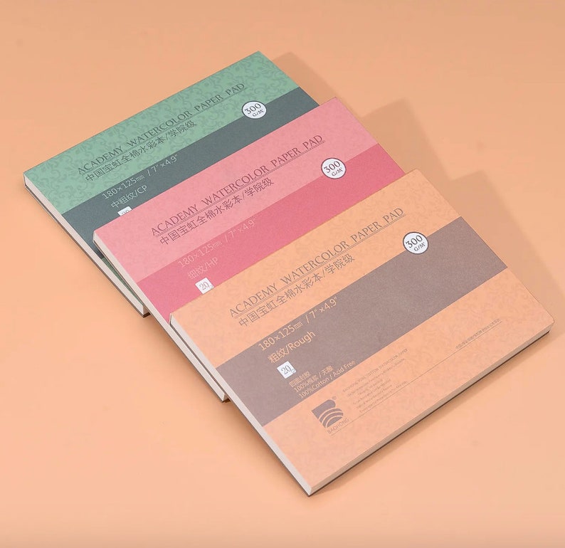 May include: Three watercolor paper pads in different colors and textures. The pads are labeled "ACADEMY WATERCOLOR PAPER PAD" and have the text "中国宝虹全棉水彩本/学院级" in Chinese. The pads are 180 x 125 mm (7 x 4.9 inches) and have different textures: "中粗纹/CP" (medium grain), "细纹/HP" (fine grain), and "粗纹/Rough" (rough grain).