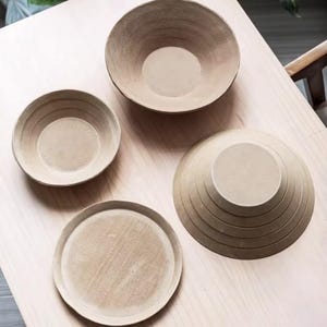 May include: Four beige ceramic bowls and plates of varying sizes are arranged on a light wood surface. The bowls have a rounded shape, while the plate is flat. The items are likely for serving food or as decorative pieces.
