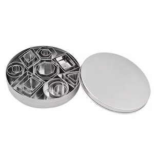 Puede incluir: Un conjunto de cortadores de galletas de acero inoxidable en varias formas geométricas, incluyendo círculos, cuadrados, triángulos y octágonos. Los cortadores están cuidadosamente dispuestos dentro de una lata redonda plateada con tapa.