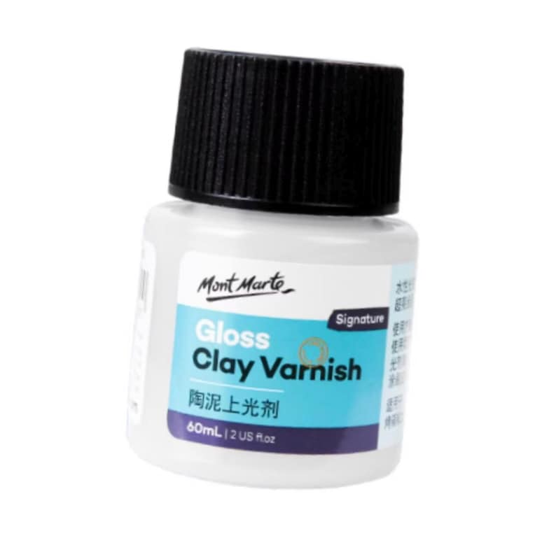 Puede incluir: Una botella blanca de barniz de arcilla brillante Mont Marte con tapa negra. La etiqueta presenta el texto "Gloss Clay Varnish" en azul y blanco, junto con el nombre de la marca y una etiqueta "Signature". La botella contiene 60 ml.