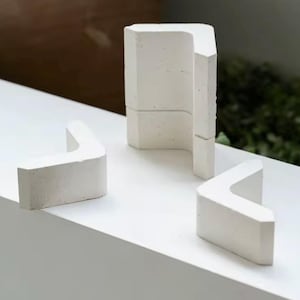 Pode incluir: Três esculturas geométricas de concreto branco. Duas têm formato de L, e uma é um bloco retangular com um interior curvo. Os objetos estão sobre uma superfície branca, com vegetação ao fundo.