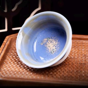 May include: A ceramic tea bowl with a light blue interior and a white and tan rim. The bowl has a glossy finish and is set on a wooden tray. The interior has a speckled design.