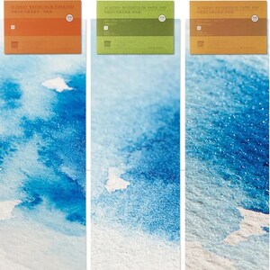 May include: Three watercolor paper pads with different grain textures: fine, medium, and coarse. Each pad has a label with the text "ACADEMY WATERCOLOR PAPER PAD" and the size of the paper in inches and centimeters. The pads are arranged from left to right in order of increasing grain size.