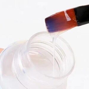 Puede incluir: Un l&iacute;quido transparente y claro se vierte de un recipiente de pl&aacute;stico transparente con un pincel. El pincel tiene un mango negro y una cabeza de pincel con tonos naranja, rojo y morado. El l&iacute;quido fluye del pincel al recipiente.