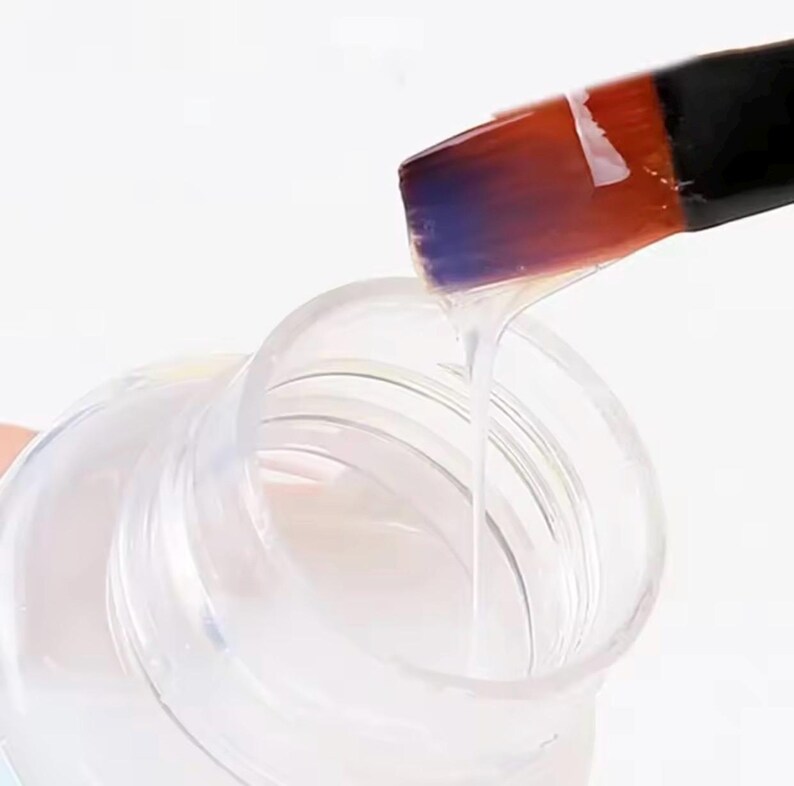 Puede incluir: Un l&iacute;quido transparente y claro se vierte de un recipiente de pl&aacute;stico transparente con un pincel. El pincel tiene un mango negro y una cabeza de pincel con tonos naranja, rojo y morado. El l&iacute;quido fluye del pincel al recipiente.