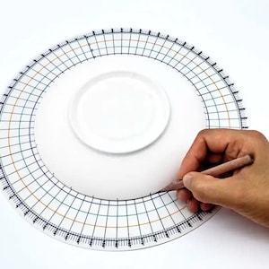 May include: A white ceramic plate with a black and orange grid pattern around the edge. A hand is holding a pencil and drawing a line on the plate.