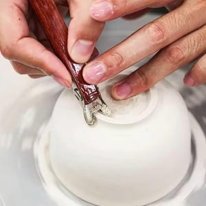 Pode incluir: Uma mão usando uma ferramenta de madeira para alisar a borda de uma tigela de cerâmica branca.