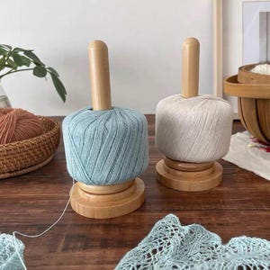 Könnte beinhalten: Zwei Garnhalter aus Holz, einer mit hellblauem Garn und der andere mit cremefarbenem Garn. Jeder hat einen zylindrischen Sockel und einen vertikalen Holzdübel. Das Garn ist ordentlich aufgewickelt, bereit für Strick- oder Häkelprojekte.