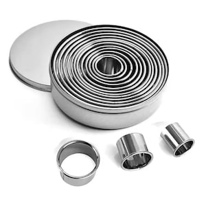 May include: A set of stainless steel round cookie cutters in various sizes, neatly stacked inside a round tin. Three individual cutters are placed nearby. The cutters are designed for baking and crafting.