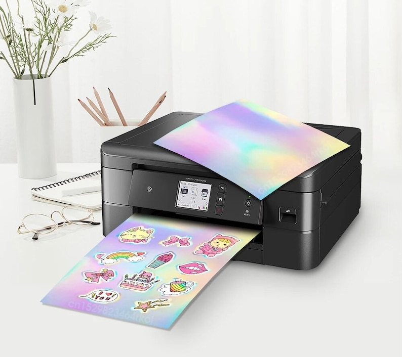 May include: A black inkjet printer printing holographic stickers with various cartoon designs. The stickers feature images of a cat, a bow, a cake, and other cute illustrations. A sheet of holographic paper rests on top.