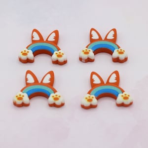 Puede incluir: Cuatro coloridos dijes de resina con forma de arcoíris con trasero de perro, patas y orejas. El arcoíris es naranja con rayas azules y verdes. Las patas son blancas con almohadillas naranjas.