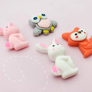 以下が含まれることがあります： ピンクの豚、白いウサギ、オレンジ色のキツネ、灰色のフクロウなど、カラフルな漫画の動物のフィギュアのコレクション。 フィギュアは滑らかな素材でできており、薄いピンク色の表面に配置されています。