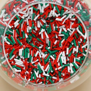 May include: A clear glass bowl filled with colorful sprinkles. The sprinkles are a mix of red, white, and green, resembling a festive holiday theme. The sprinkles are small, cylindrical shapes, perfect for decorating baked goods.