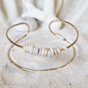 Può includere: Un bracciale a polsino in metallo dorato con una fila di perline di conchiglia bianche e beige. Il bracciale è ambientato su uno sfondo sabbioso, evocando un tema costiero. Il gioiello è semplice ed elegante.