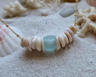 May include: A delicate necklace featuring a strand of small, white seashells and a single, light blue sea glass bead. The necklace is strung on a fine gold chain, resting on a bed of sand with seashells and coral.