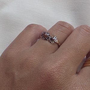 May include: A thin gold band ring featuring two small, textured, shell-like ornaments side by side. The ornaments have a dark, oxidized finish with white highlights, creating a detailed, radial, fan-shaped pattern on the surface of the ring.
