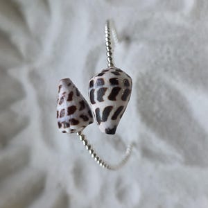May include: A silver ring with two cone-shaped seashells, patterned with black and brown spots. The ring is set against a white, textured background, evoking a natural, seaside theme.