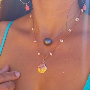 May include: A woman wearing a turquoise swimsuit with a layered necklace featuring a large black pearl, a small seashell pendant, and a strand of white seashells. The necklace is accented with a gold chain. She is also wearing a pair of hoop earrings with a pink and white seashell charm.