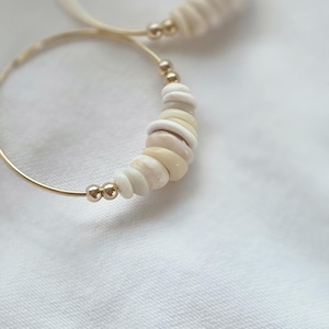 May include: A pair of gold hoop earrings featuring a series of small, smooth, off-white and cream-colored disc beads interspersed with tiny metallic gold spheres. The beads are arranged along the lower curve of each thin, circular gold wire hoop.