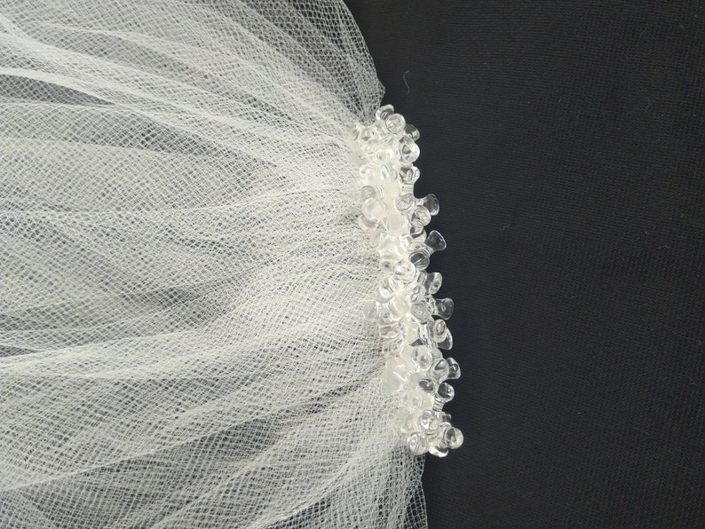May include: White tulle bridal veil with a beaded edge. The veil is made of soft, delicate fabric and has a simple, elegant design.
