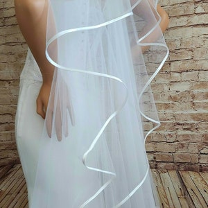 May include: A white bridal veil with a flowing design. The veil is trimmed with a white satin ribbon that creates a wavy, decorative edge. The veil is sheer and drapes over a white wedding dress.