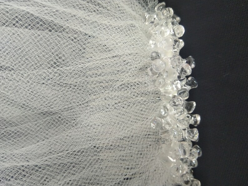 May include: White tulle fabric with a beaded trim. The trim is made of clear plastic beads.