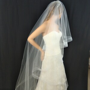 May include: A white bridal veil with a single layer of tulle and a scalloped edge. The veil is worn over a white wedding dress.