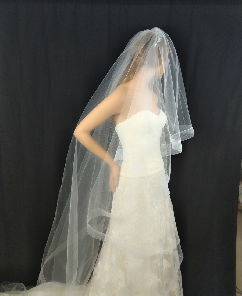 May include: A white bridal veil with a single layer of tulle and a scalloped edge. The veil is worn over a white wedding dress.