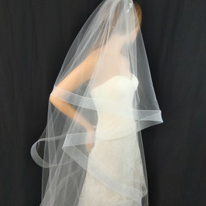 May include: A white bridal veil with a single layer of tulle and a delicate beaded embellishment. The veil is draped over a white wedding dress.