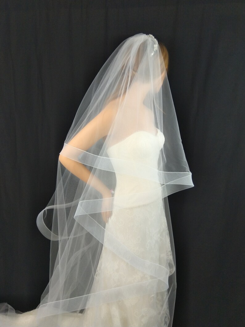 May include: A white bridal veil with a single layer of tulle and a delicate beaded embellishment. The veil is draped over a white wedding dress.
